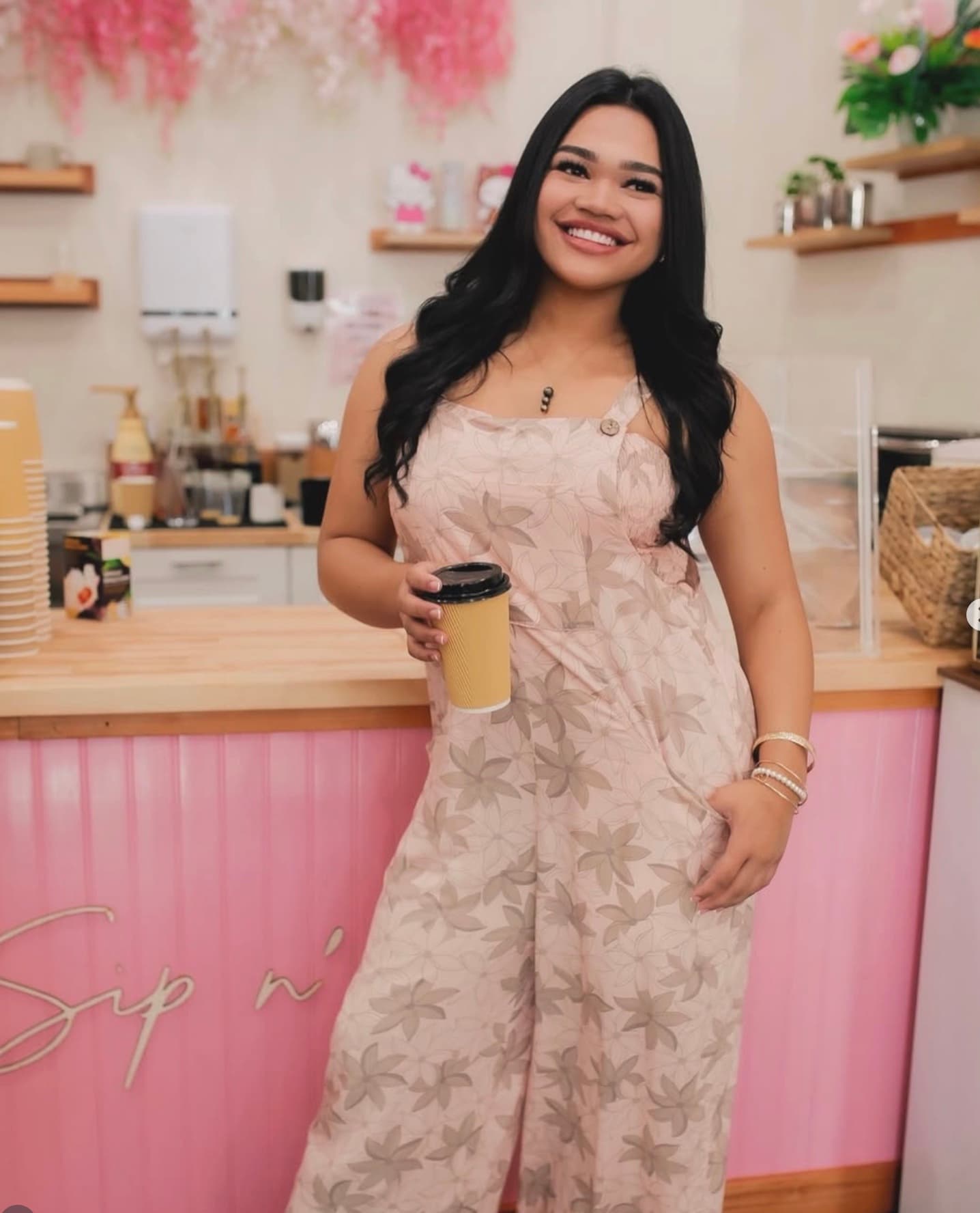A barista at the pink scalloped counter of Sip N' Pretty, holding a hot latte, wearing a floral jumpsuit.