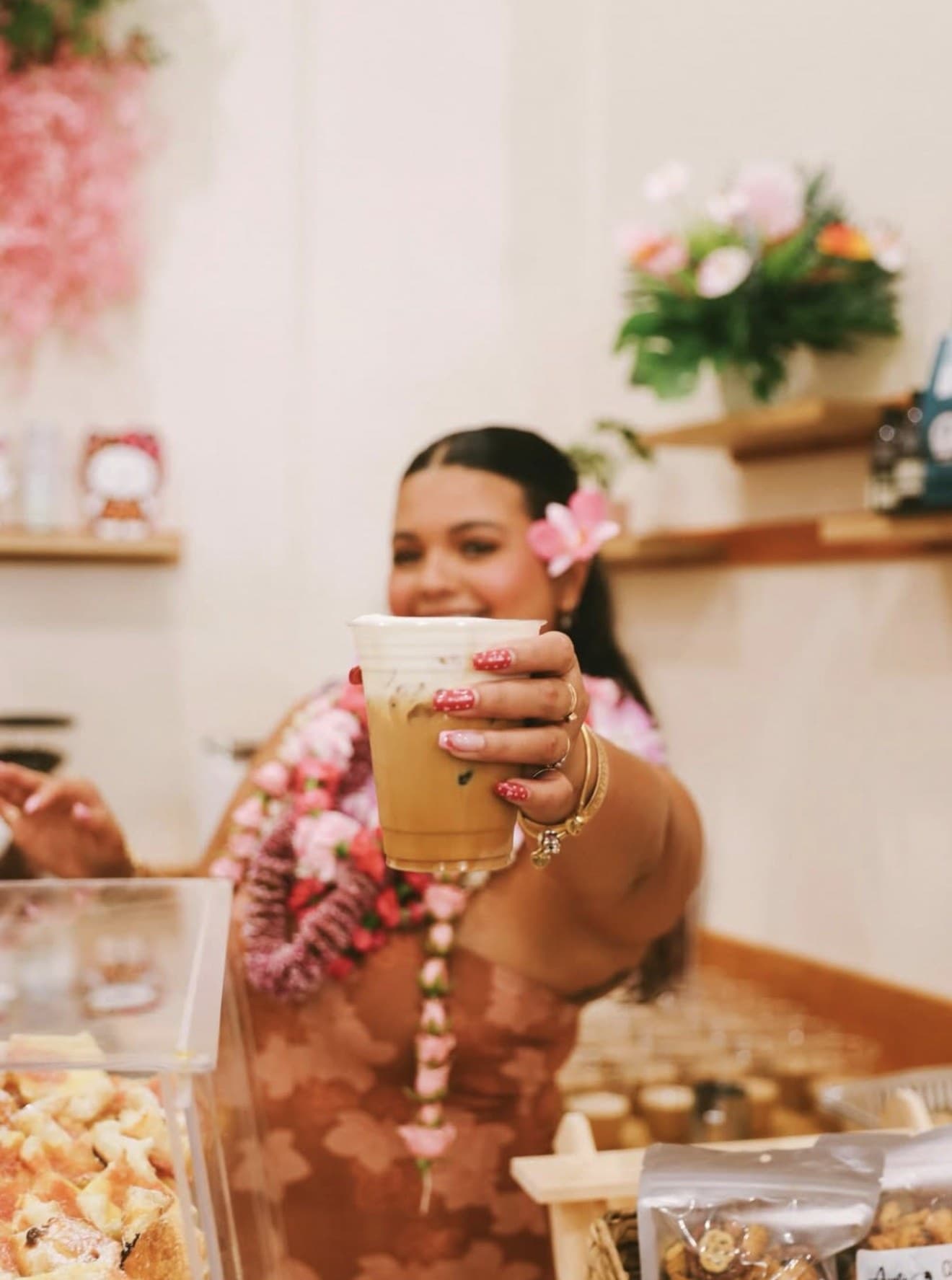 A barista at Sip N' Pretty in Hilo holding up an iced latte, fresh plumeria leis around her neck, flowers behind the bar.