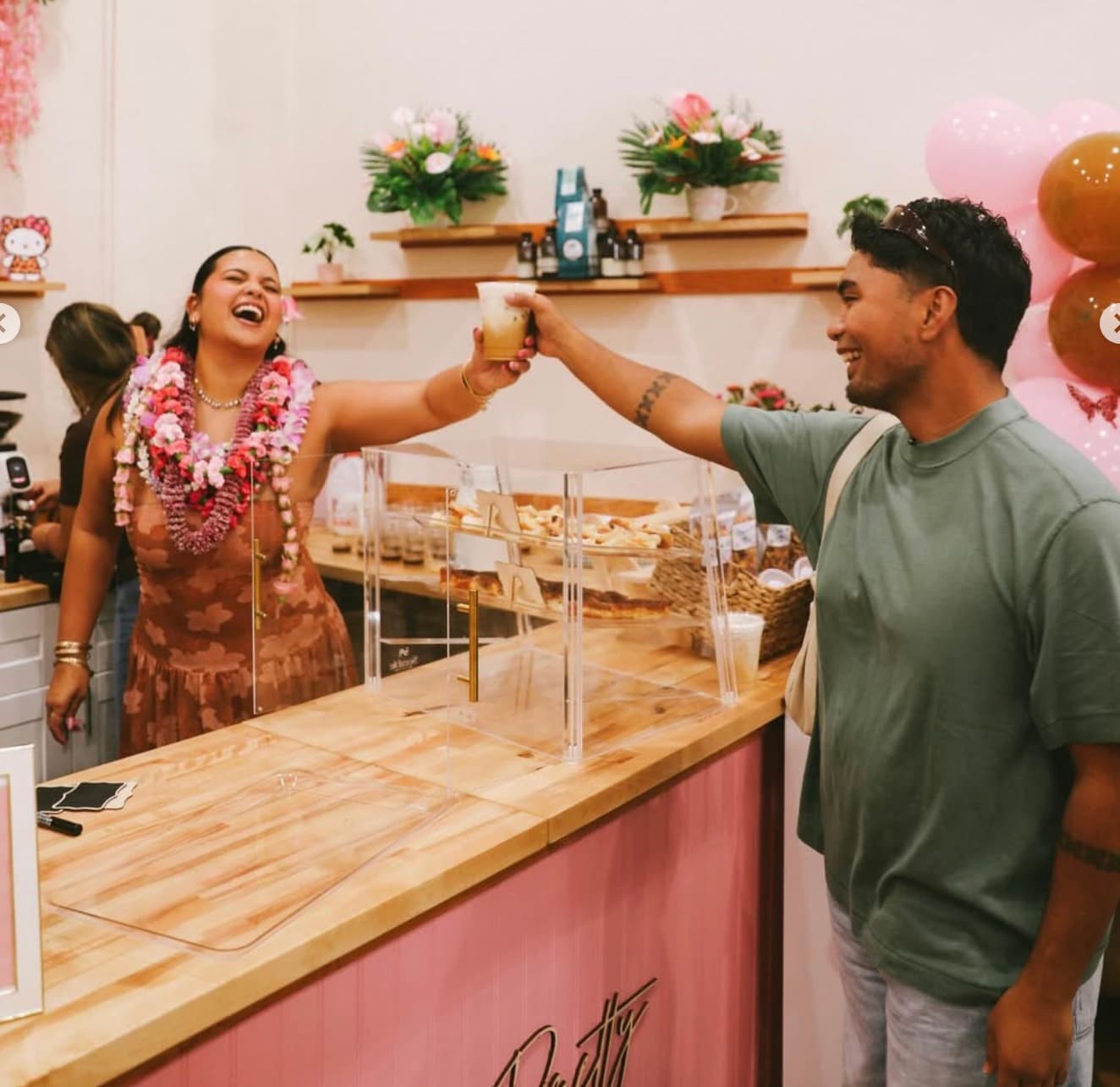 Barista and customer fist-bumping over the bar at Sip N' Pretty, donut case and pink balloons.