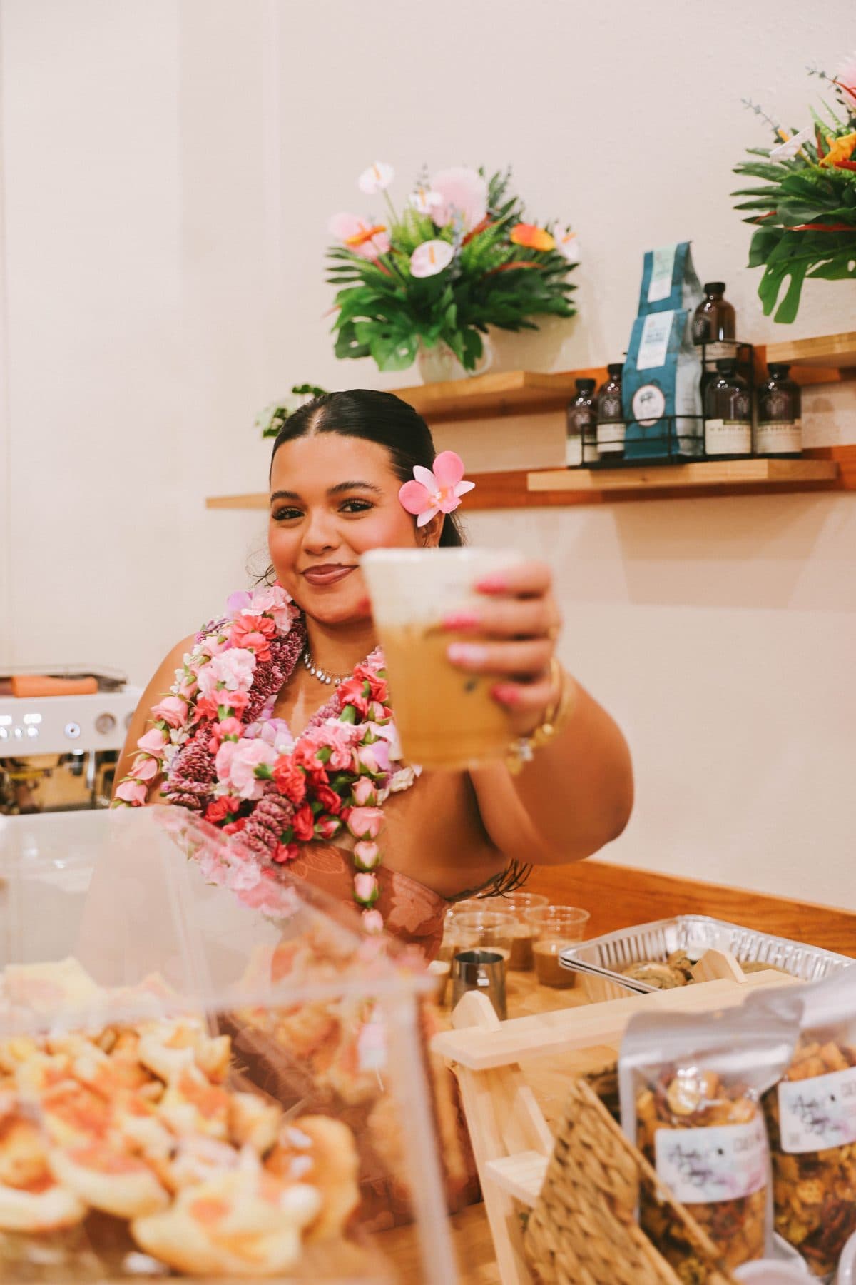 Ani at the Sip n' Pretty counter in Hilo, holding up an iced latte, draped in fresh flower lei.